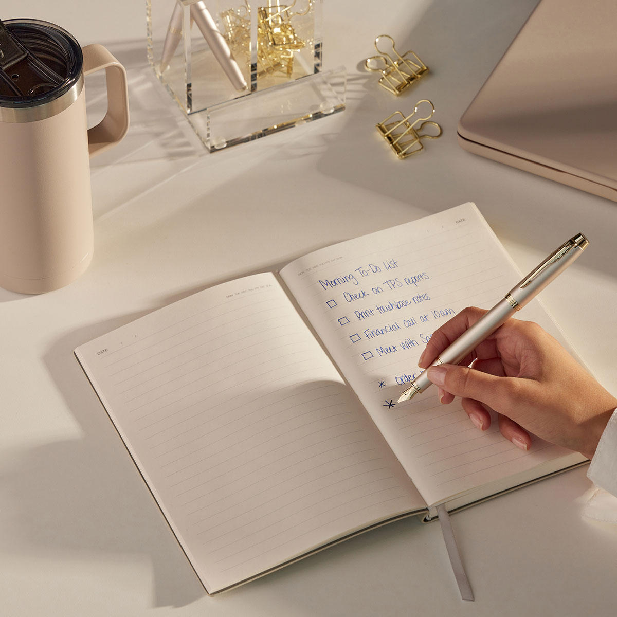 Osoba zapisuje notatki w otwartym notesie eleganckim piórem, na biurku leżą akcesoria biurowe i kubek.